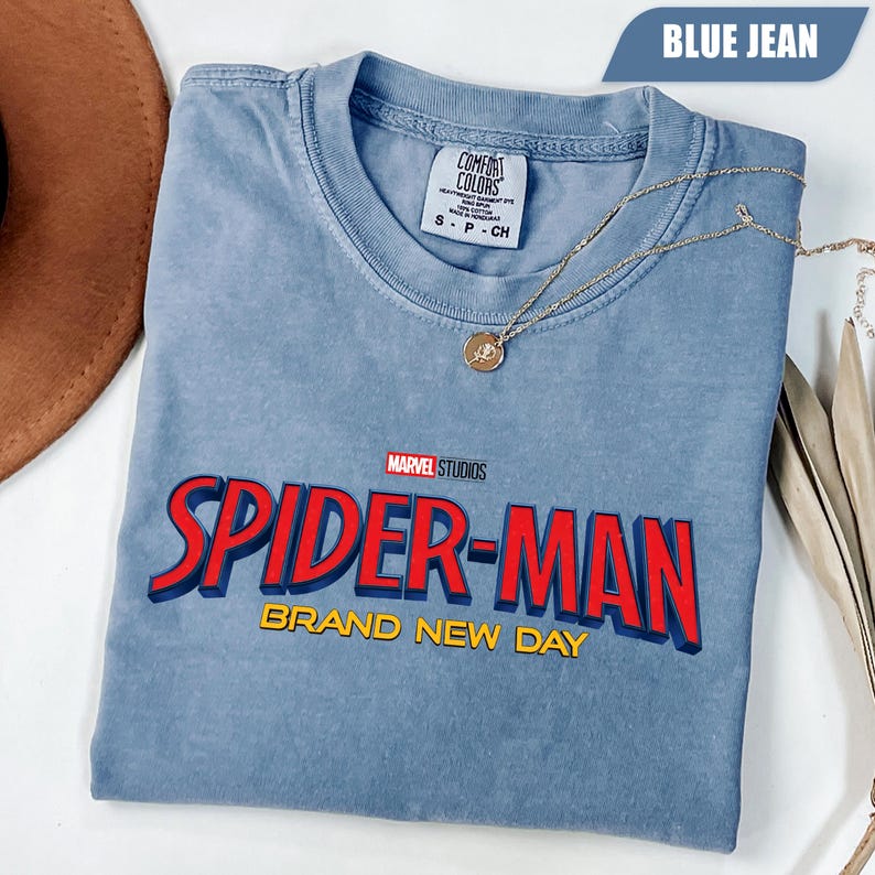 Może przedstawiać: Niebieska koszulka w kolorze dżinsu z logo Spider-Man z Marvel Studios w kolorze czerwonym i ż&oacute;łtym. Koszulka ma napis "BRAND NEW DAY" pod logo. Złoty naszyjnik z wisiorkiem jest na koszulce.
