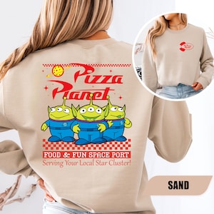 Puede incluir: Sudadera de color arena con un dise&ntilde;o de Pizza Planet. La parte trasera muestra el logotipo de Pizza Planet y tres personajes alien&iacute;genas verdes. El texto dice "FOOD & FUN SPACE PORT Serving Your Local Star Cluster!". Un logotipo m&aacute;s peque&ntilde;o de Pizza Planet est&aacute; en la parte delantera.