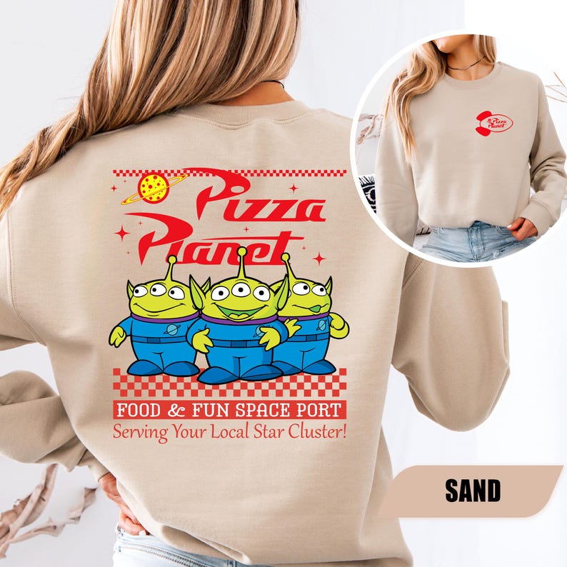 Puede incluir: Sudadera de color arena con un dise&ntilde;o de Pizza Planet. La parte trasera muestra el logotipo de Pizza Planet y tres personajes alien&iacute;genas verdes. El texto dice "FOOD & FUN SPACE PORT Serving Your Local Star Cluster!". Un logotipo m&aacute;s peque&ntilde;o de Pizza Planet est&aacute; en la parte delantera.