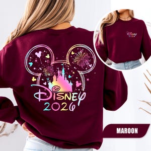 Puede incluir: Sudadera granate con un dise&ntilde;o colorido de Disney que incluye un castillo, fuegos artificiales y el texto "Disney 2026". El dise&ntilde;o tiene forma de orejas de Mickey Mouse. Una versi&oacute;n m&aacute;s peque&ntilde;a del dise&ntilde;o est&aacute; en la parte delantera. La palabra "MAROON" est&aacute; en la parte inferior.