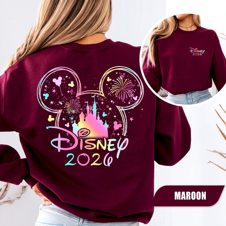 Puede incluir: Sudadera granate con un dise&ntilde;o colorido de Disney que incluye un castillo, fuegos artificiales y el texto "Disney 2026". El dise&ntilde;o tiene forma de orejas de Mickey Mouse. Una versi&oacute;n m&aacute;s peque&ntilde;a del dise&ntilde;o est&aacute; en la parte delantera. La palabra "MAROON" est&aacute; en la parte inferior.