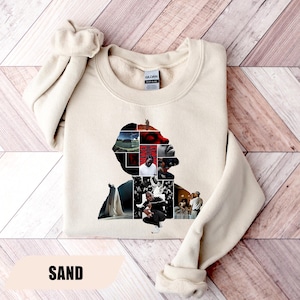 May include: Sand-colored sweatshirt featuring a graphic silhouette filled with various images. The design includes a collage of photos within the shape of a person's profile. The word "SAND" is printed at the bottom of the image.