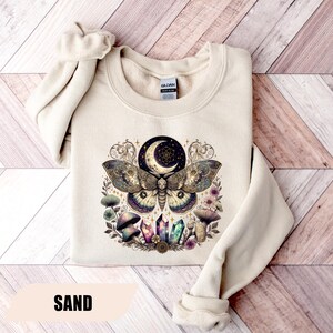May include: Sand-colored sweatshirt featuring a detailed graphic of a moth with a crescent moon, crystals, and mushrooms. The design incorporates shades of gold, purple, and green. The word "SAND" is printed at the bottom of the sweatshirt.