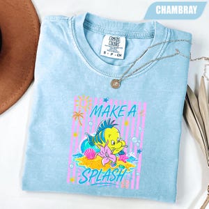 May include: Light blue t-shirt with a graphic of a yellow fish, a pink flower, and the words "MAKE A SPLASH". The shirt has a crew neck and short sleeves. A silver necklace with a pendant is on top of the shirt.
