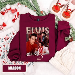 May include: Maroon sweatshirt featuring a collage of Elvis Presley images and his signature. The word "ELVIS" is displayed in a sparkling font. The sweatshirt is surrounded by holiday decorations, including candy canes and a white knit hat.