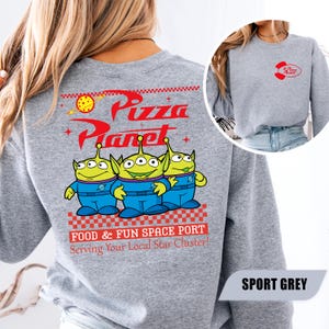 Puede incluir: Sudadera gris deportivo con el logo de Pizza Planet y tres personajes alien&iacute;genas verdes. El dise&ntilde;o trasero incluye las palabras "Pizza Planet" en rojo, con la frase "FOOD & FUN SPACE PORT Serving Your Local Star Cluster!"