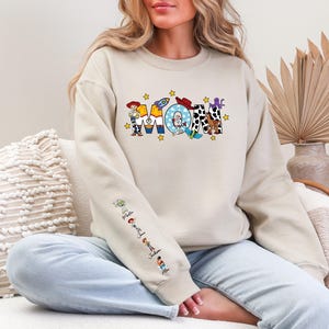 Op de afbeelding: Een beige sweatshirt met het woord "MOM" in kleurrijke, cartoonachtige letters. De letters zijn versierd met verschillende personages en afbeeldingen. De mouw heeft extra stripfiguren. De sweatshirt is een casual, comfortabele stijl.
