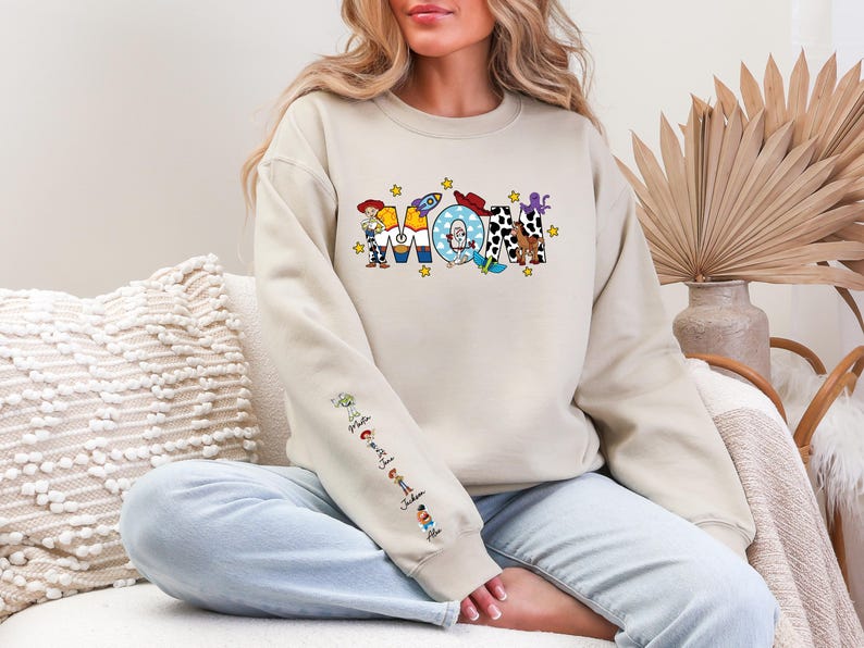 Op de afbeelding: Een beige sweatshirt met het woord "MOM" in kleurrijke, cartoonachtige letters. De letters zijn versierd met verschillende personages en afbeeldingen. De mouw heeft extra stripfiguren. De sweatshirt is een casual, comfortabele stijl.