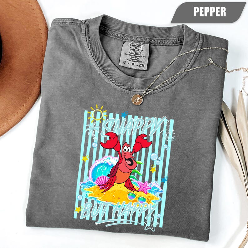 May include: Pepper-colored Comfort Colors t-shirt featuring a graphic of a red crab on a sandy beach with the words "Snappy But Happy". The design includes blue stripes, a wave, and a starfish. A silver necklace is draped across the shirt.