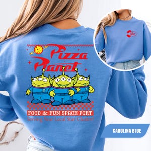 Puede incluir: Sudadera azul Carolina con el logo de Pizza Planet y tres personajes alien&iacute;genas verdes. El dise&ntilde;o trasero incluye el texto "Pizza Planet" y "FOOD & FUN SPACE PORT Serving Your Local Star Cluster!". Un logo m&aacute;s peque&ntilde;o est&aacute; en el frente.