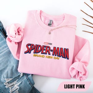 Puede incluir: Sudadera rosa claro de Spider-Man con el logo de Marvel Studios. El texto "SPIDER-MAN" es rojo y azul, con "BRAND NEW DAY" en dorado. Un collar dorado con un colgante est&aacute; sobre la sudadera.