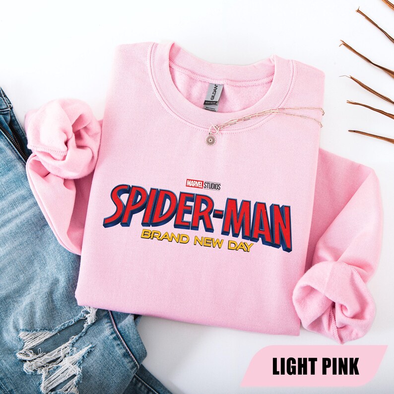 Puede incluir: Sudadera rosa claro de Spider-Man con el logo de Marvel Studios. El texto "SPIDER-MAN" es rojo y azul, con "BRAND NEW DAY" en dorado. Un collar dorado con un colgante est&aacute; sobre la sudadera.
