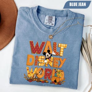 Puede incluir: Una camiseta color azul vaquero con el texto WALT DISNEY WORLD en letras grandes y estampadas. Mickey Mouse, Chip, Chop y Pluto aparecen ilustrados entre hojas de otoño y una calabaza. Un collar de tono dorado descansa sobre el cuello.