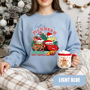 May include: Light blue sweatshirt featuring a graphic of Lightning McQueen and other Cars characters in Santa hats and Christmas trees. The text "Lightning McQueen" and "Merry Christmas" are also visible. A gingerbread man mug is held in the hand.