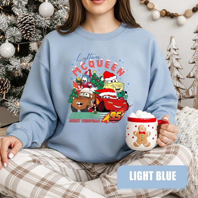 May include: Light blue sweatshirt featuring a graphic of Lightning McQueen and other Cars characters in Santa hats and Christmas trees. The text "Lightning McQueen" and "Merry Christmas" are also visible. A gingerbread man mug is held in the hand.
