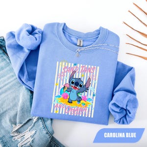 May include: A Carolina blue crewneck sweatshirt featuring a graphic of the character Stitch holding an ice cream cone. The design includes the text SAND, TANS AND OHANA, along with illustrations of a sun, seashells, and a starfish on a striped background.