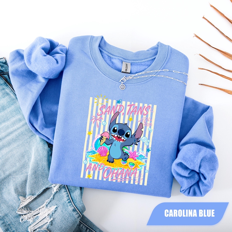 May include: A Carolina blue crewneck sweatshirt featuring a graphic of the character Stitch holding an ice cream cone. The design includes the text SAND, TANS AND OHANA, along with illustrations of a sun, seashells, and a starfish on a striped background.