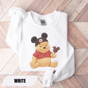 Pu&ograve; includere: Felpa bianca con un'illustrazione di Winnie the Pooh con un cappello di Topolino e che tiene in mano una caramella a forma di Topolino. La felpa ha una camicia rossa sotto e la parola "WHITE" &egrave; in basso.