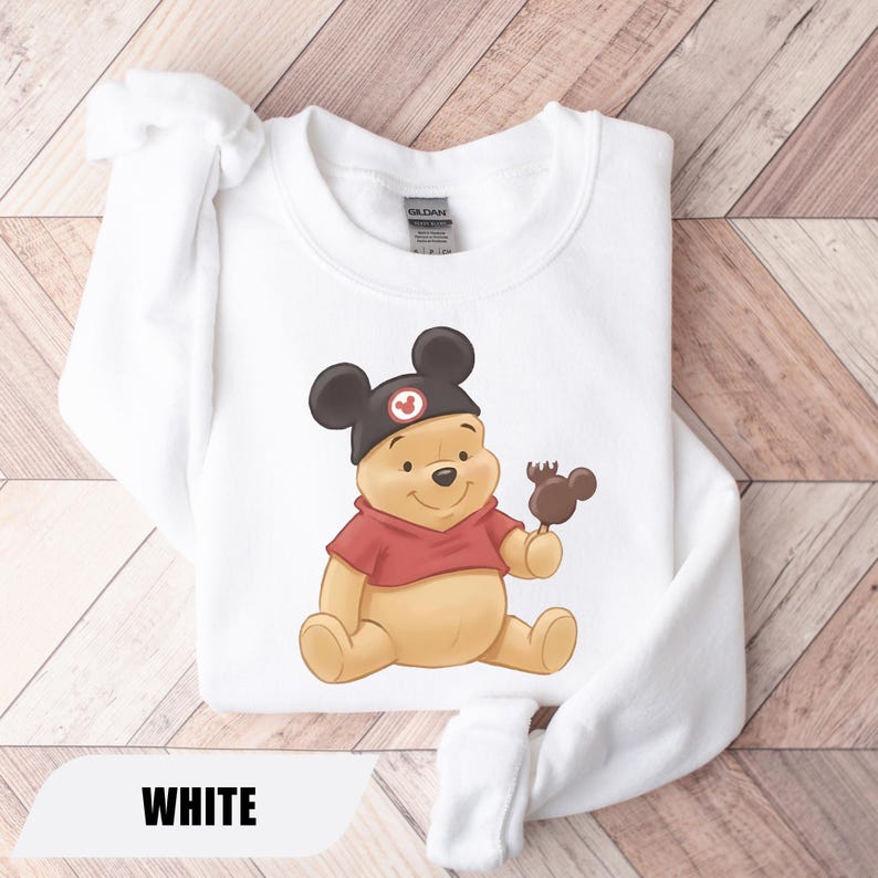 Pu&ograve; includere: Felpa bianca con un'illustrazione di Winnie the Pooh con un cappello di Topolino e che tiene in mano una caramella a forma di Topolino. La felpa ha una camicia rossa sotto e la parola "WHITE" &egrave; in basso.