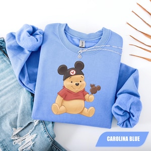 Pu&ograve; includere: Felpa blu Carolina con un personaggio dei cartoni animati di Winnie the Pooh con un cappello di Topolino e che tiene in mano una caramella a forma di Topolino. La felpa ha un girocollo e maniche lunghe. Il testo "CAROLINA BLUE" &egrave; in un rettangolo blu in basso.