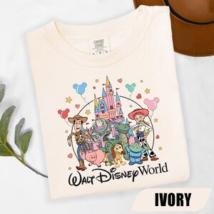Puede incluir: Camiseta de color marfil con un gr&aacute;fico de personajes de Disney frente a un castillo, con las palabras "Walt Disney World" debajo. La camiseta incluye personajes como Woody, Buzz Lightyear y Jessie. La palabra "IVORY" est&aacute; impresa en la parte inferior.