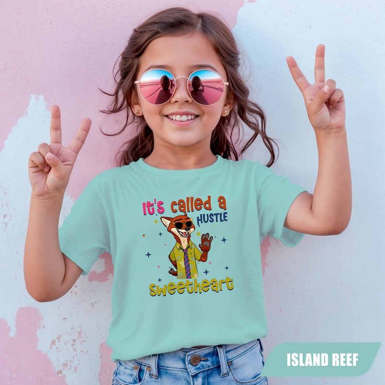 K&ouml;nnte beinhalten: Ein hellblaues T-Shirt mit dem Text "It's called a hustle sweetheart" und einer Cartoon-Fuchsfigur. Das T-Shirt wird von einem Kind getragen, das eine runde Sonnenbrille tr&auml;gt und mit den Fingern ein Friedenszeichen macht.