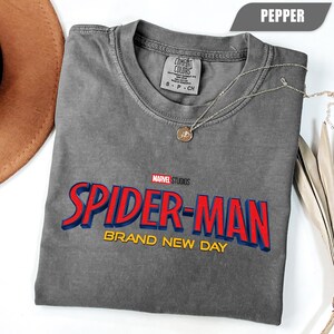 Może przedstawiać: Szara koszulka Spider-Man w kolorze pieprzu z czerwonym i niebieskim logo oraz napisem "BRAND NEW DAY". Koszulka, marki Comfort Colors, ma okrągły dekolt i kr&oacute;tkie rękawy. Srebrny naszyjnik z wisiorkiem jest na koszulce.