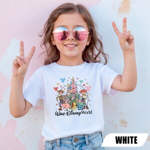 Puede incluir: Una camiseta blanca con un gr&aacute;fico colorido de personajes de Disney y un castillo, con el texto "Walt Disney World" en escritura negra. La camiseta la lleva una persona que lleva gafas de sol y hace el signo de la paz.