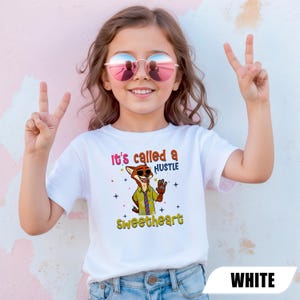 K&ouml;nnte beinhalten: Ein wei&szlig;es T-Shirt mit einem Cartoon-Fuchs-Charakter mit Sonnenbrille und dem Text "It's called a HUSTLE Sweetheart". Das T-Shirt wird von einer jungen Person getragen, die eine rosa Sonnenbrille tr&auml;gt und ein Friedenszeichen macht.