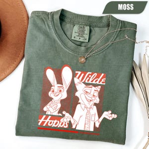 Puede incluir: Camiseta verde musgo con un gr&aacute;fico de Judy Hopps y Nick Wilde de Zootopia. El dise&ntilde;o incluye las palabras "Wilde" y "Hopps" en rojo. La camiseta tiene cuello redondo y est&aacute; hecha de un material suave. Tambi&eacute;n se ve un collar plateado con un colgante.