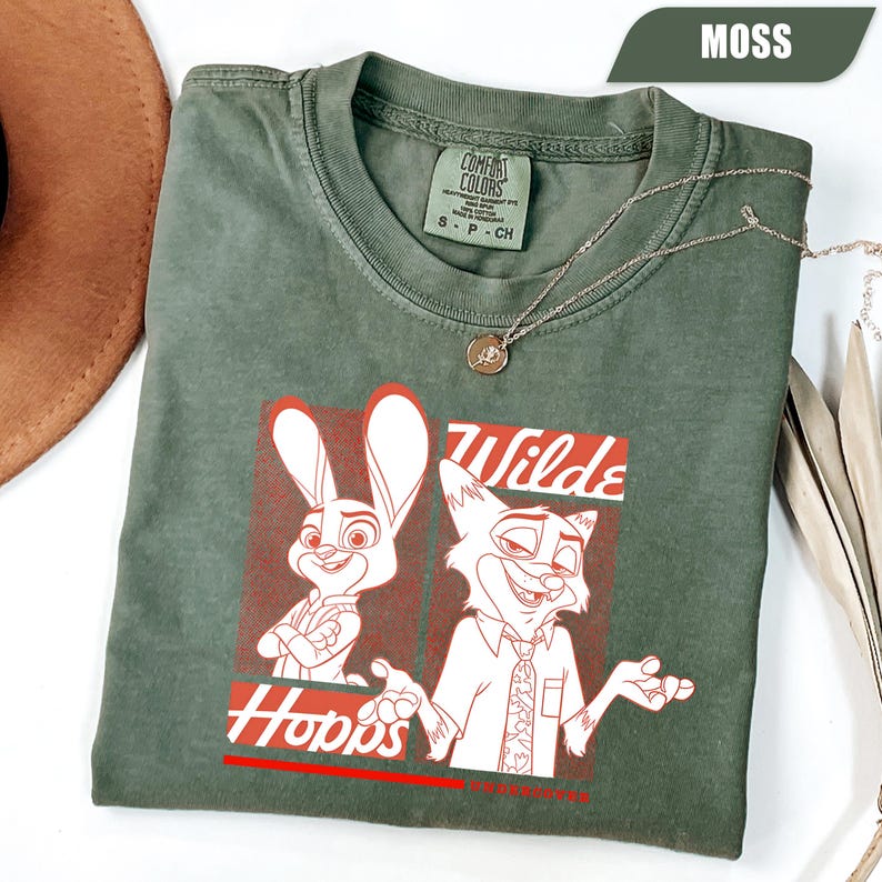 Puede incluir: Camiseta verde musgo con un gr&aacute;fico de Judy Hopps y Nick Wilde de Zootopia. El dise&ntilde;o incluye las palabras "Wilde" y "Hopps" en rojo. La camiseta tiene cuello redondo y est&aacute; hecha de un material suave. Tambi&eacute;n se ve un collar plateado con un colgante.