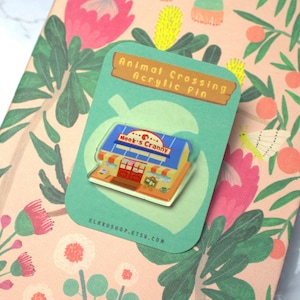May include: An Animal Crossing acrylic pin of Nook's Cranny, displayed on a floral notebook. The pin is on a teal card with the text "Animal Crossing Acrylic Pin". The notebook has a pink cover with floral and leaf designs.