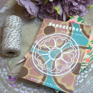 May include: A clear, circular suncatcher with a white floral design, layered over a patterned card with the text "Suncatcher The White Lotus...". A spool of twine and decorative flowers are also in the image.