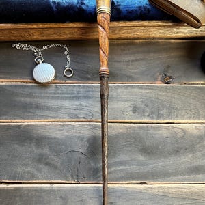 Puede incluir: Una varita de madera hecha a mano con un mango de latón y un diseño en espiral. La varita es de color marrón oscuro con un patrón de vetas de madera más claro. Un reloj de bolsillo plateado con una cadena es visible en el fondo.