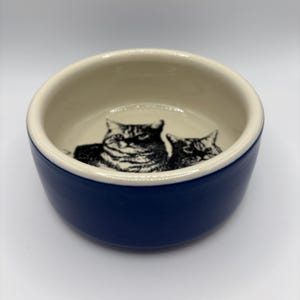 May include: A navy blue ceramic pet bowl with a cream interior. The inside of the bowl features a black and white illustration of two cats. The bowl has a rounded shape and is suitable for cats or small dogs.