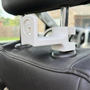 May include: A white 3D-printed plastic car headrest hook featuring a prominent letter G on the front face. The hook is designed to attach securely to the metal support posts of a vehicle headrest, providing a sturdy hanger for personal items.