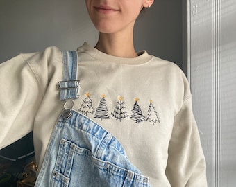 Può includere: Una felpa beige con una fila di cinque alberi di Natale ricamati in nero con stelle dorate in cima. La felpa è indossata sopra una salopette di jeans blu chiaro.