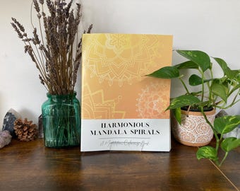 May include: A coloring book titled "Harmonious Mandala Spirals" with a yellow and white mandala design. The book is on a wooden surface, next to a green glass vase with dried lavender and a potted plant.