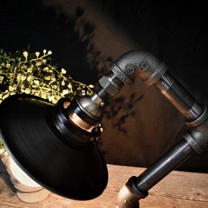 Puede incluir: Una lámpara de escritorio de estilo industrial negro con una pantalla de metal negro y un brazo de estilo tubería. La lámpara está hecha de metal negro y tiene un aspecto industrial vintage.