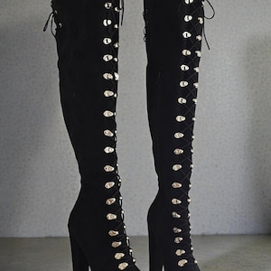 Könnte beinhalten: Ein Paar schwarze Overknee-Stiefel mit hohen Absätzen und Plateausohlen. Die Stiefel haben ein Schnürdesign mit goldfarbenen Ösen und eine weiche, Wildleder-ähnliche Textur. Die Stiefel sind für einen kühnen, modischen Look konzipiert.