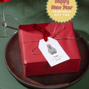 May include: A wrapped red gift box with a tag that says "Happy New Year Printable Gift Tag". The tag features a watercolor illustration of a capybara wearing a party hat. The gift is on a wooden plate.