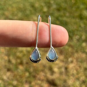 May include: A pair of silver teardrop earrings with a blue-gray stone. The earrings have a beaded border and a long, slender hook. The earrings are displayed against a blurred green background.