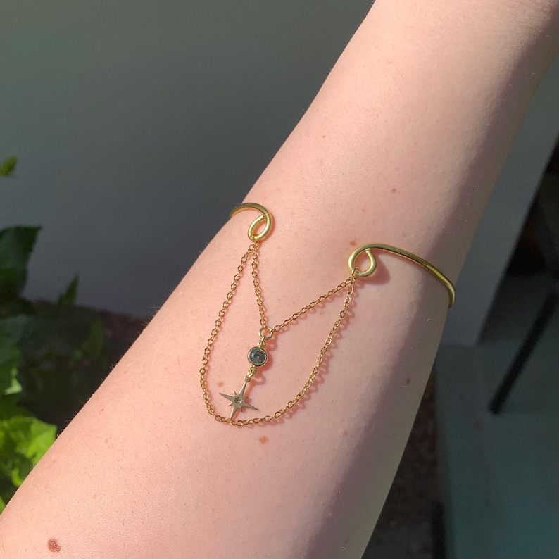 May include: A gold chain bracelet with a dangling star charm and a small round stone. The bracelet is worn on a person's arm.