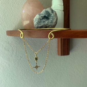 May include: A gold necklace with a star charm and a green gemstone pendant. The necklace is hanging from a gold wire that is attached to a wooden shelf.