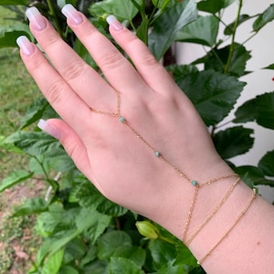 May include: A gold chain bracelet with green beads, designed to be worn on the hand and wrist. The bracelet has a delicate chain and is adorned with small, round green beads.
