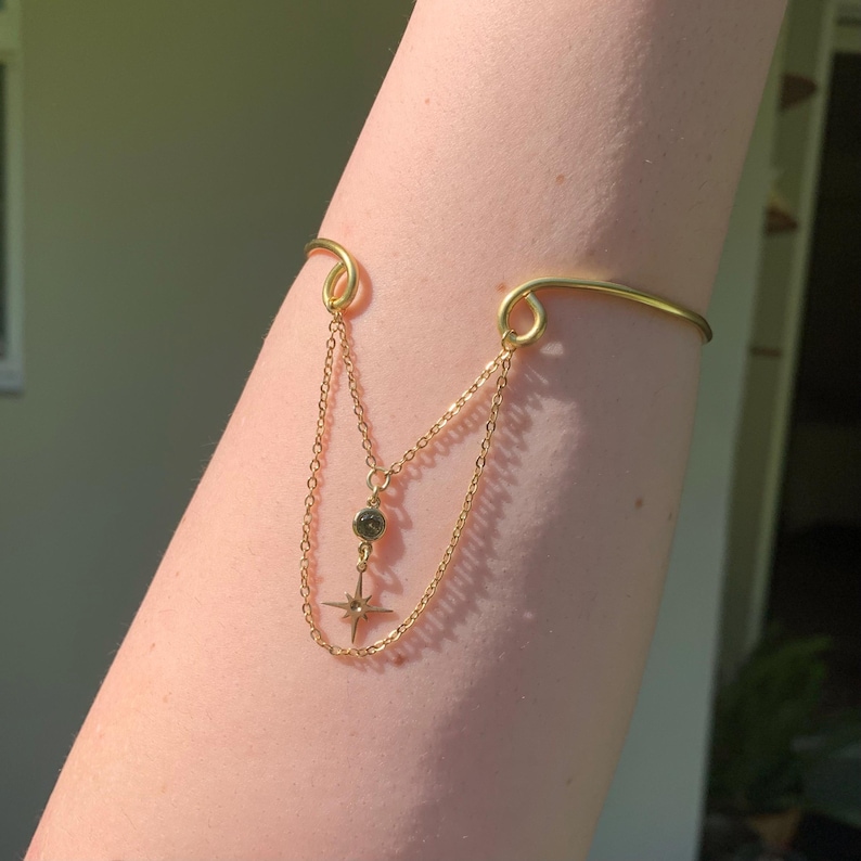 May include: A gold arm cuff with two chains and a star charm. The star charm has a small, round, clear stone in the center.
