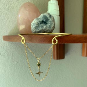 May include: A gold choker necklace with a dangling star charm and a green gemstone. The necklace is on a wooden shelf.