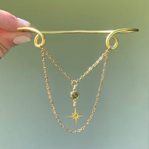 May include: A gold chain necklace with a green gemstone pendant and a star charm. The necklace is attached to a gold bar with two loops at the ends.
