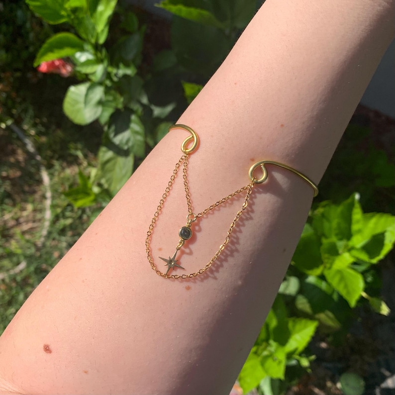 May include: A gold arm cuff with a dangling chain and a star charm. The chain has a small black gemstone.