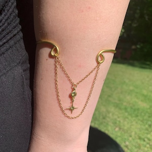 May include: A gold arm cuff with a dangling chain and a star charm with a green gemstone. The cuff is worn on a person's arm.
